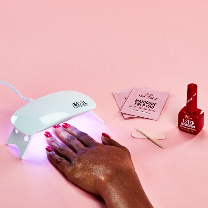 Hand under Ardell LED lamp curing Venetian Red gel polish, with manicure tools and prep pads on a pink background.