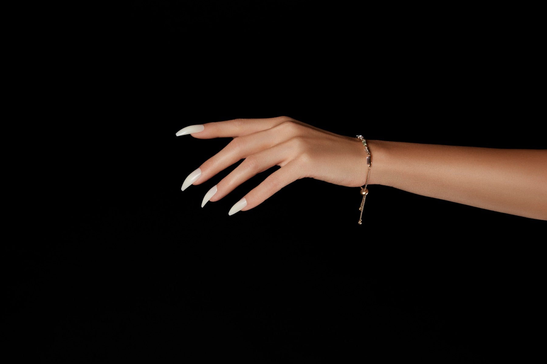 A gracefully posed hand featuring glossy natural ballerina long nails with a delicate gold bracelet against a black background.
