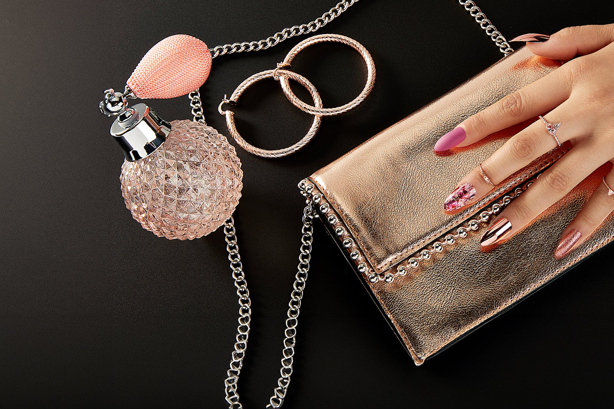 Holiday party-ready beauty close-up with pink and metallic nail art, rose-gold clutch, crystal perfume bottle, and sparkling accessories.