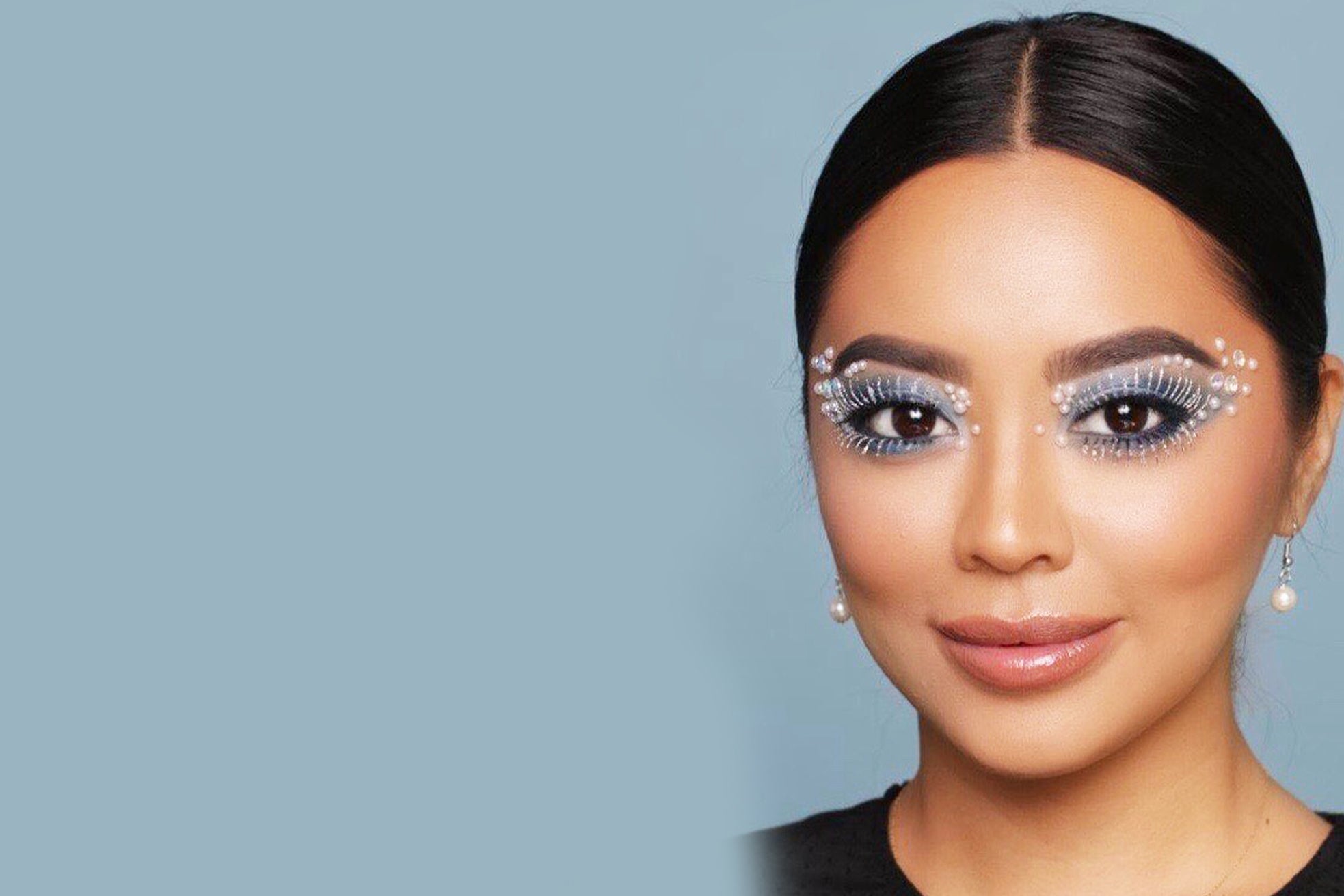 Woman with decorative eye makeup, false lashes, and pearl earrings against a dark background