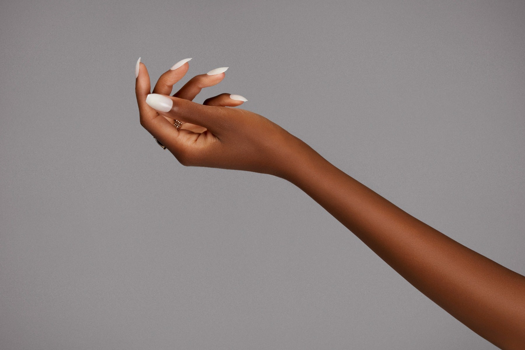 Curved almond white nails on extended hand with minimal gold rings, set against a gray backdrop.