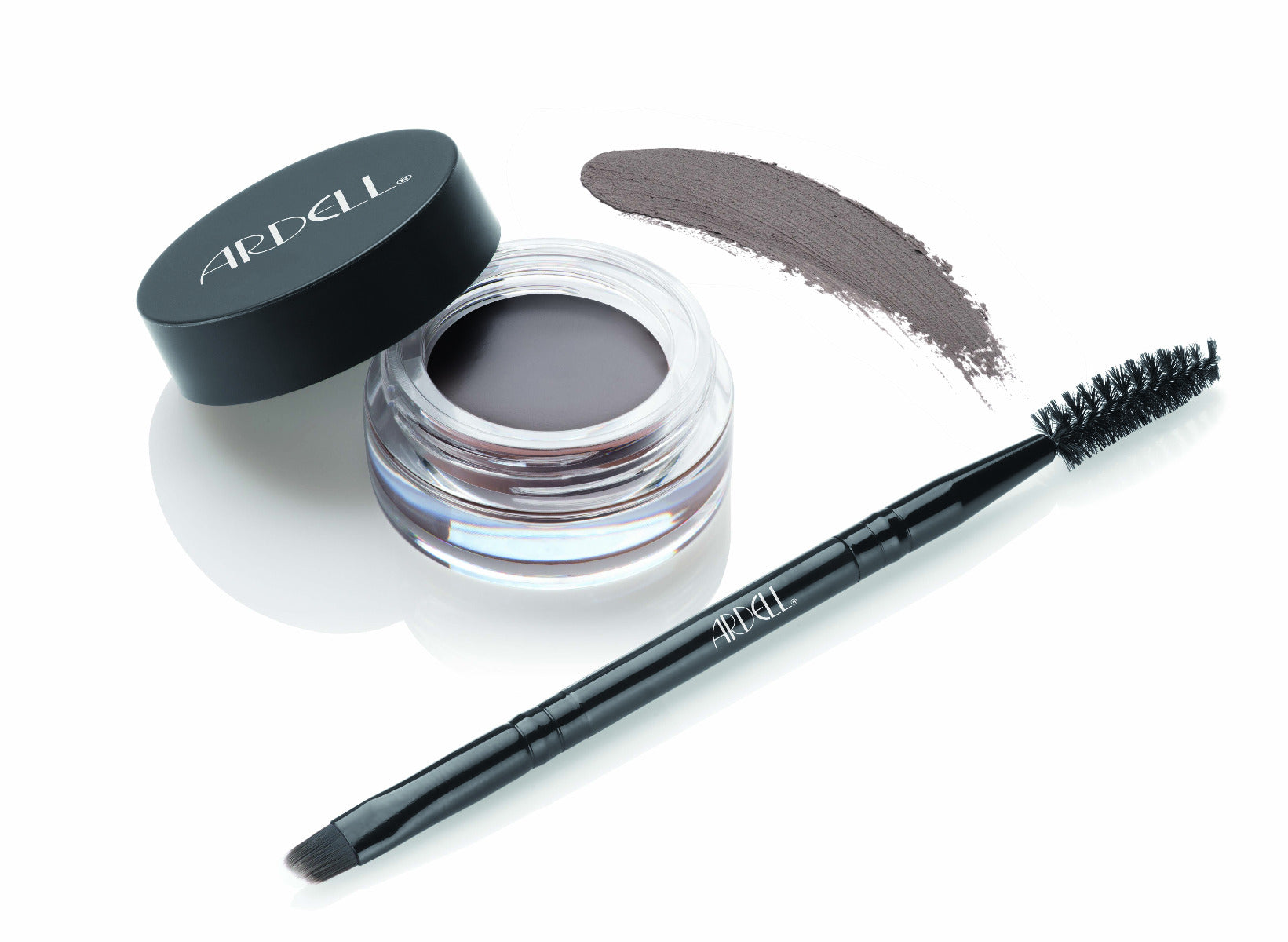 Uncapped container of Ardell Brow Pomade alongside brush/spoolie tool with pomade swatched onto background to show color